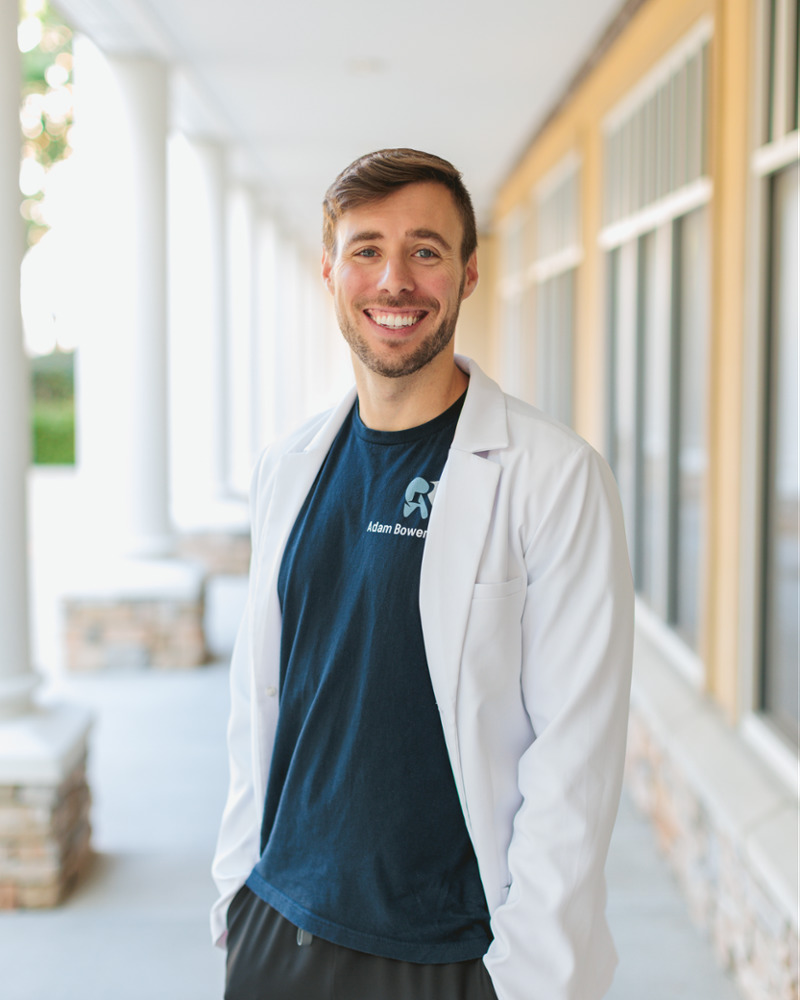 Dr. Adam Bowers, Durham dentist, smiling outdoors in a white lab coat and branded scrub shirt