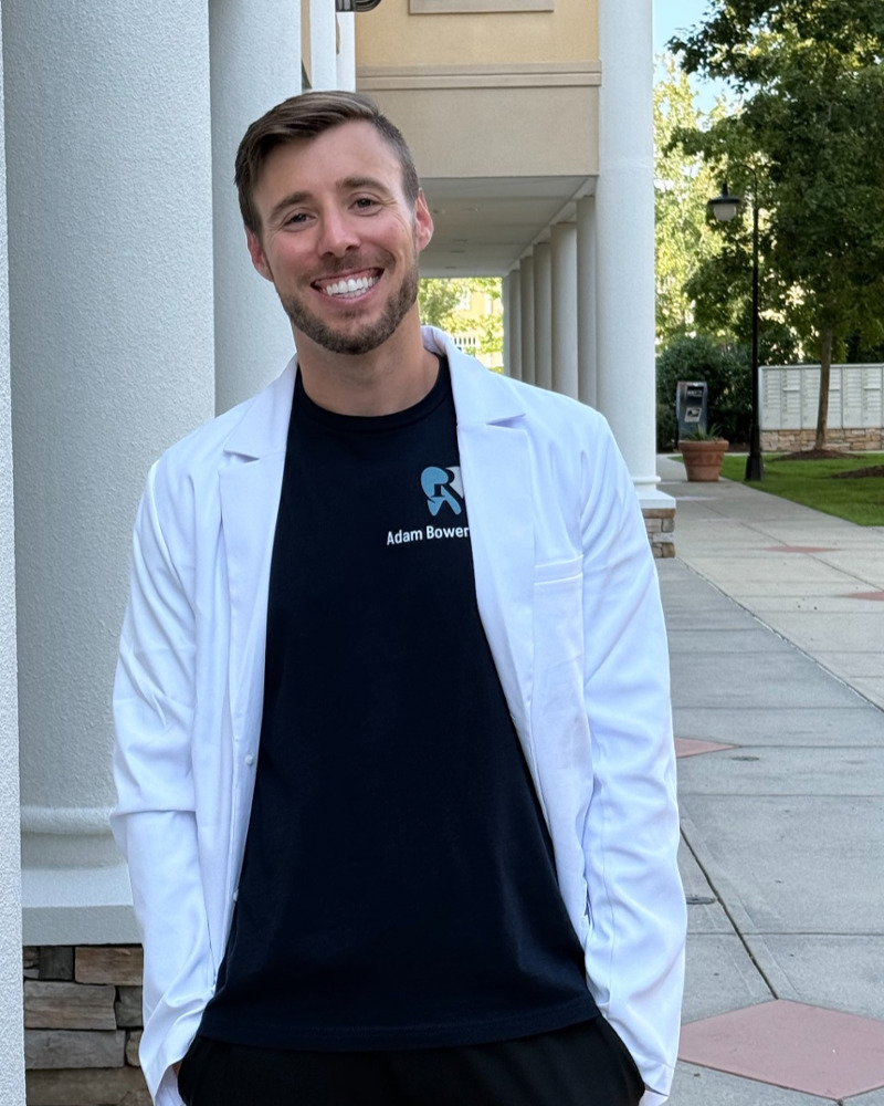 Dr. Adam Bowers, Durham dentist, smiling outdoors in a white lab coat and branded scrub shirt